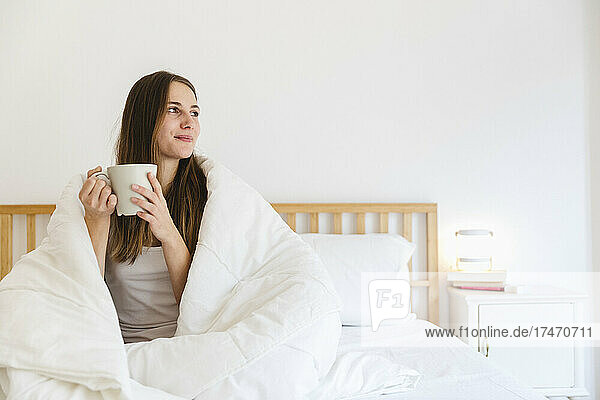 Young woman with brown hair having coffee on bed at home