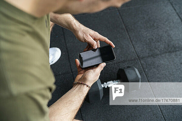 Young sportsman photographing dumbbell through smart phone in gym