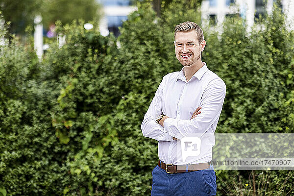 Smiling young businessman standing with arms crossed at park