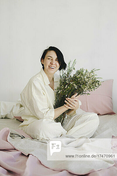 Cheerful woman with flower bouquet sitting on bed at new home
