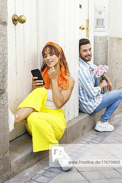 Smiling girlfriend using mobile phone while sitting at doorway with man