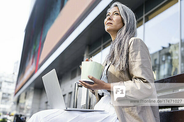 Thoughtful woman holding lunch box while sitting with laptop on bench
