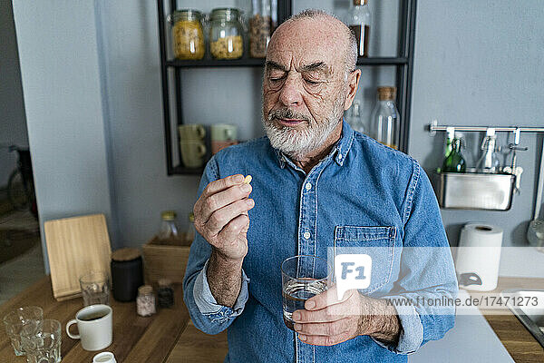 Senior man holding glass of water looking at medicine in kitchen