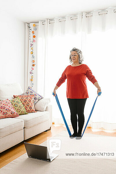 Senior woman doing gymnastic with resistance band while watching tutorial on laptop at home