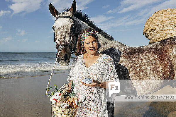 Pregnant mother with baby booties standing by horse at beach