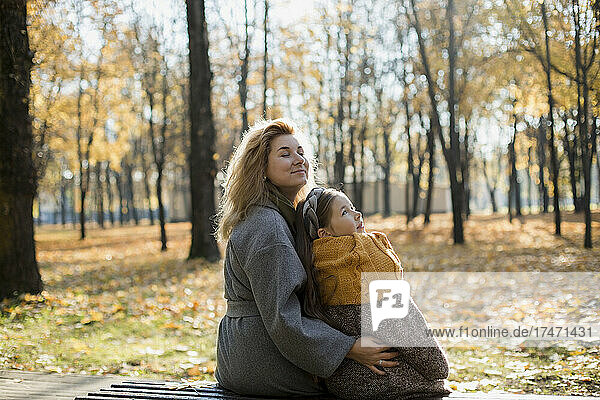 Mother with eyes closed embracing daughter on park bench