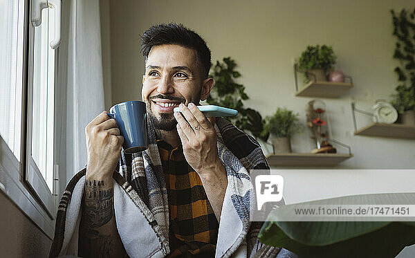 Lächelnder Mann hält Kaffeetasse in der Hand  während er am Fenster mit dem Mobiltelefon spricht