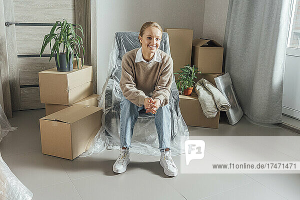 Smiling young woman sitting on armchair wrapped in plastic at home