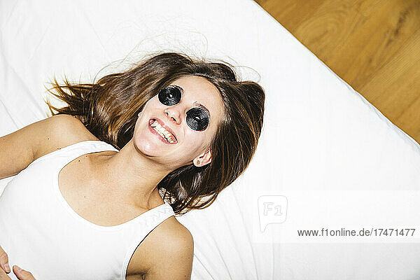 Glückliche junge Frau  die ihre Augen mit Lakritzbonbons bedeckt  während sie zu Hause im Bett liegt
