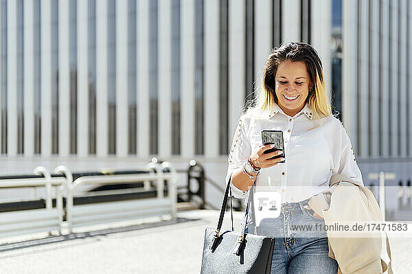 Lächelnde Freiberuflerin mit Tasche und Textnachrichten auf dem Smartphone in der Stadt