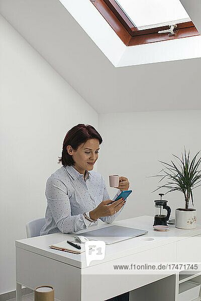 Businesswoman using smart phone while having coffee at desk in home office