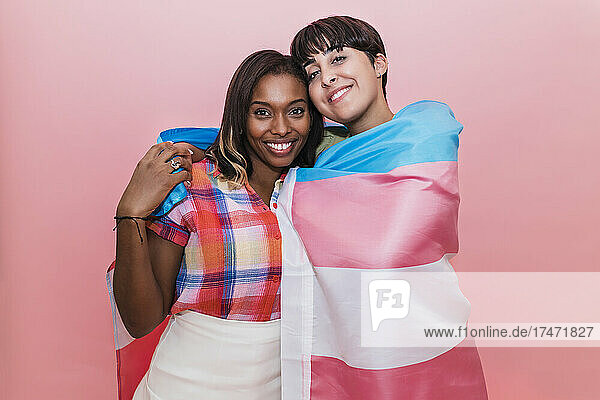 Smiling lesbian couple covered in transgender flag against pink background