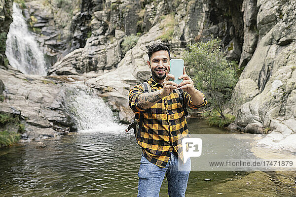 Smiling mid adult man taking selfie through smart phone by pond in forest