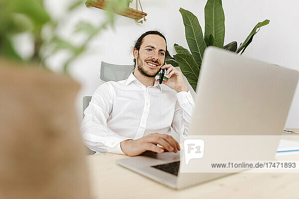 Businessman talking on mobile phone while using laptop in office