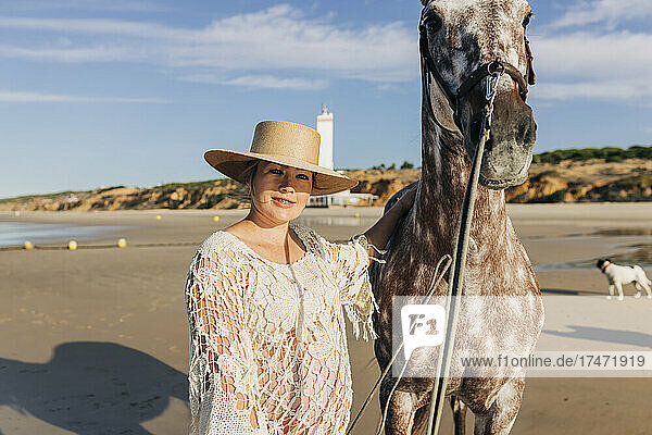 Pregnant woman wearing hat standing with horse at beach