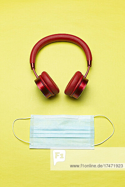 Studio shot of red wireless headphones and protective face mask lying against yellow background
