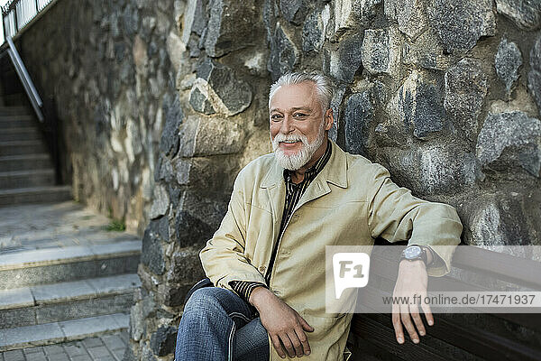Lächelnder reifer Mann sitzt auf einer Bank an einer Steinmauer