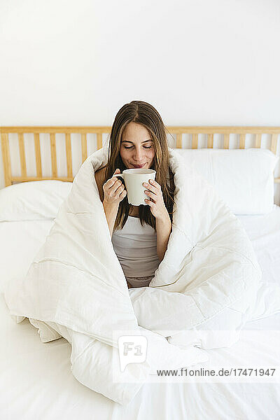 Young woman with eyes closed drinking coffee while wrapped in blanket at home