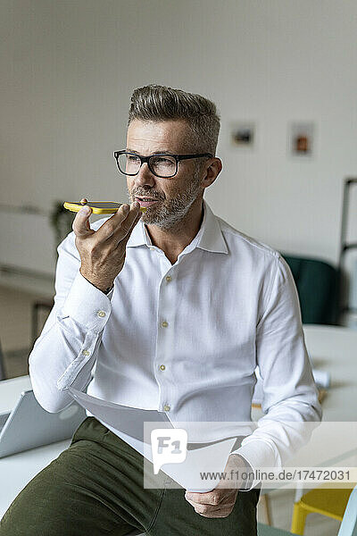 Businessman holding document while talking on smart phone in office