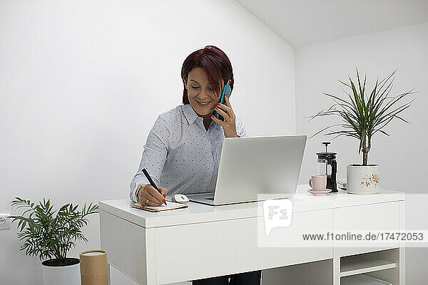 Happy female freelancer writing in note pad while talking on smart phone at home office