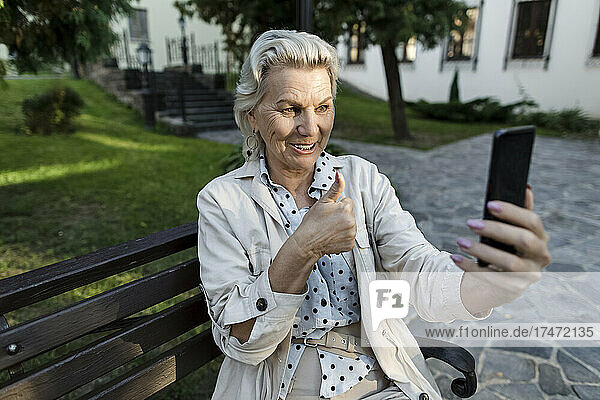 Lächelnde Frau gestikuliert während eines Videoanrufs über ihr Mobiltelefon auf der Bank mit dem Daumen nach oben