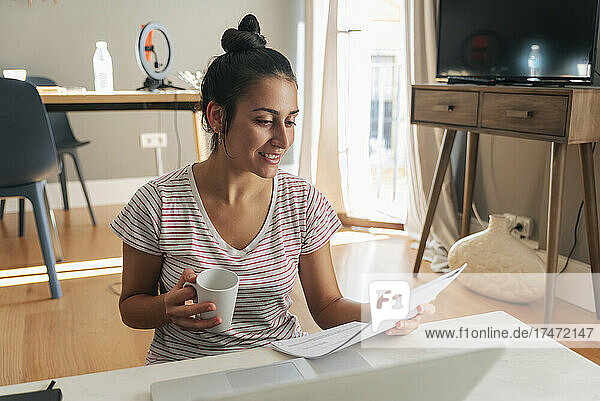 Lächelnde Geschäftsfrau mit Kaffeetasse liest Dokument im Heimbüro