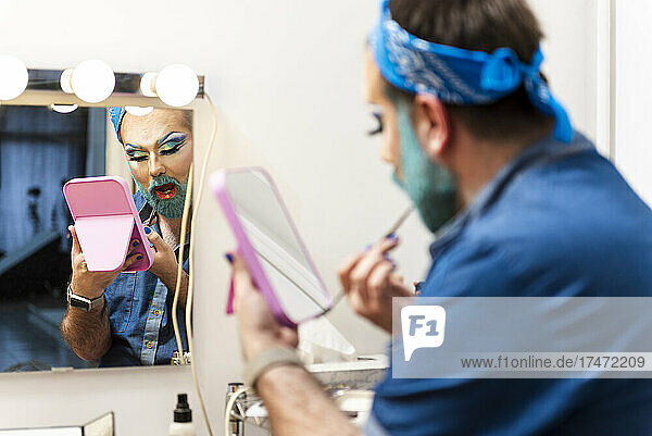Bearded man with hand mirror applying makeup in dressing room