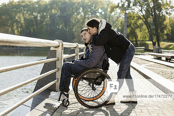 Girlfriend kissing boyfriend sitting in wheelchair at embankment