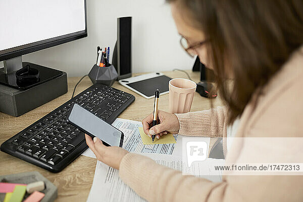 Businesswoman holding smart phone and writing while sitting at desk