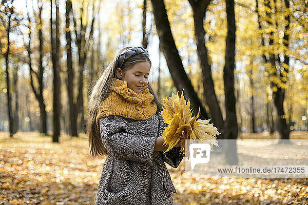 Smiling girl wearing woolen scarf holding bunch of autumn leaves in park