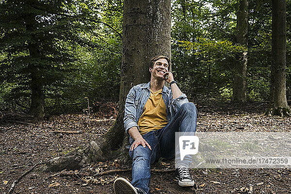Happy man with hand on chin sitting by tree trunk in forest