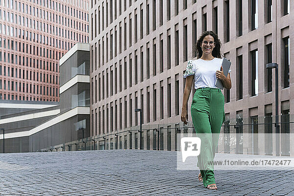 Smiling young businesswoman walking with laptop on footpath