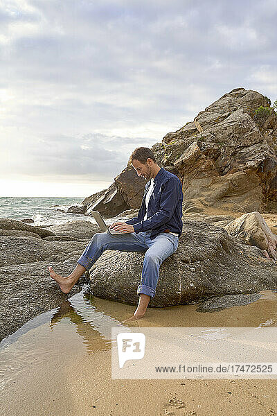 Man sitting on rock using laptop at beach