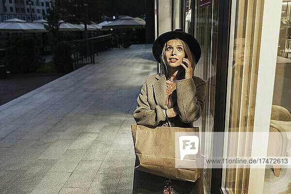 Woman talking on mobile phone by store window at dusk