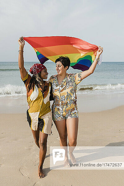Sorgloses lesbisches Paar mit Regenbogenfahne spaziert am Strand