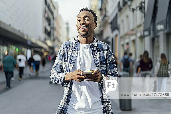 Smiling young man holding smart phone at city street