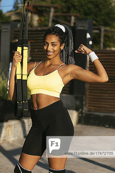 Smiling sportswoman flexing bicep while holding suspension straps