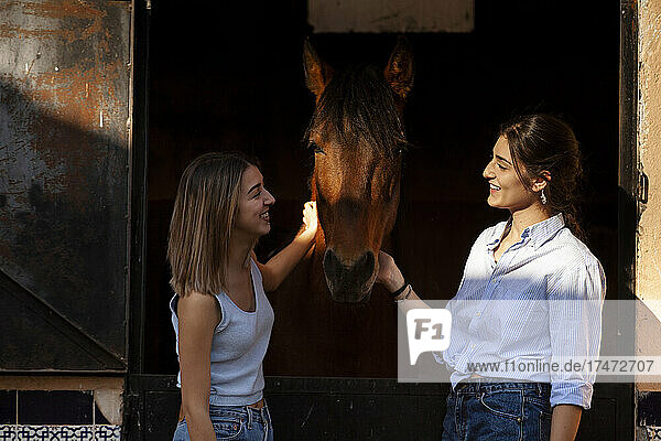 Smiling friends talking and stroking horse at stable