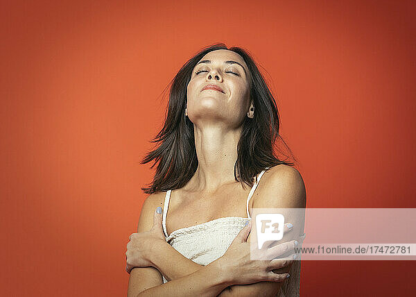 Smiling woman with eyes closed hugging self against orange background