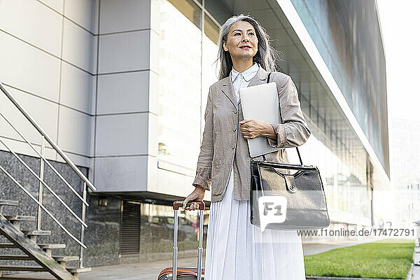 Smiling female professional with luggage and laptop standing at office park