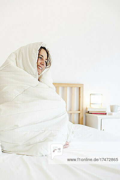 Glückliche junge Frau  die in eine Decke gehüllt ist  während sie zu Hause im Bett sitzt
