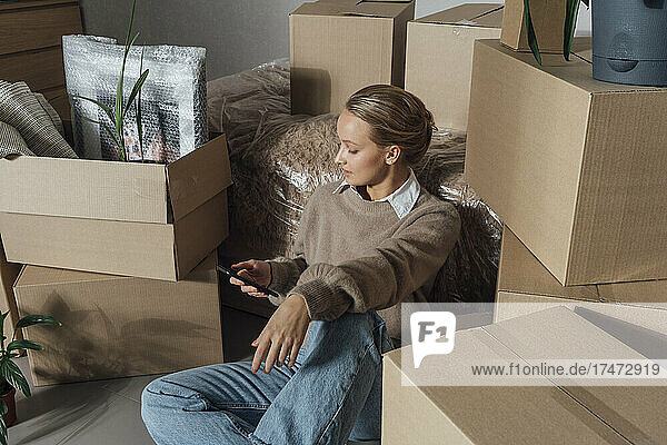Young woman using smart phone amidst cartons in new home