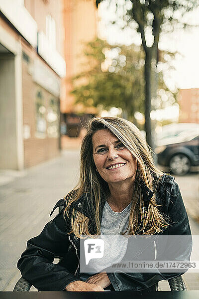 Lächelnde blonde Frau in Lederjacke sitzt im Straßencafé