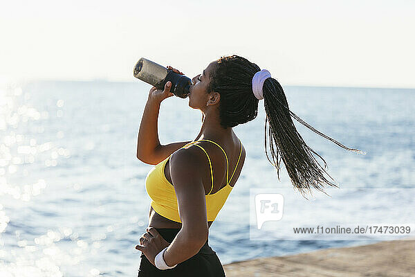 Young sportswoman drinking water while standing near sea