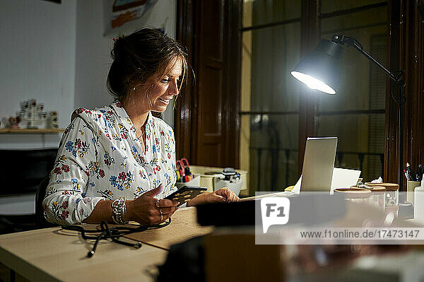 Businesswoman holding smart phone while working on laptop in office