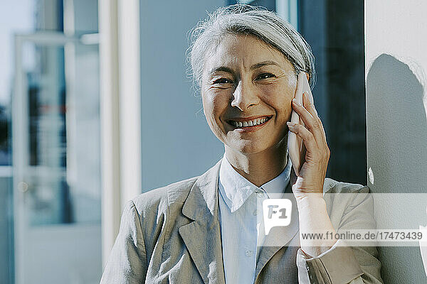 Happy mature woman talking on mobile phone by wall