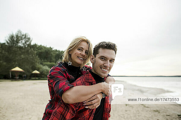 Smiling young man giving piggyback ride to blond woman at beach
