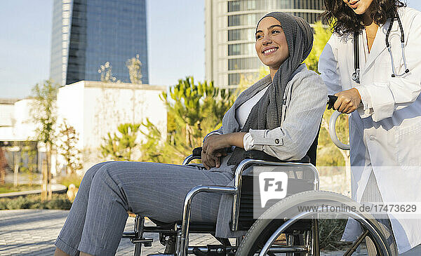 Junge Allgemeinärztin schiebt den Rollstuhl einer lächelnden behinderten Frau auf dem Fußweg