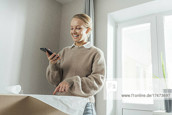 Smiling young woman with mobile phone pointing at plan on carton