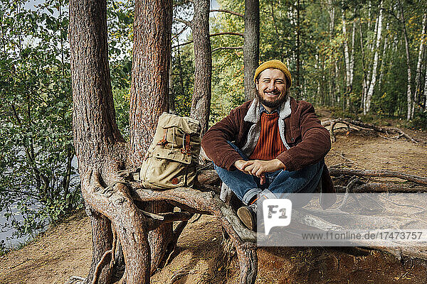 Lächelnder Mann mit Rucksack sitzt auf Baumwurzeln im Wald